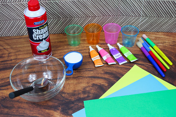 A photo of the ingredients needed to make puffy paint. These include a bowl, shaving cream, food coloring, glue, paint brushes, and colored cardstock.