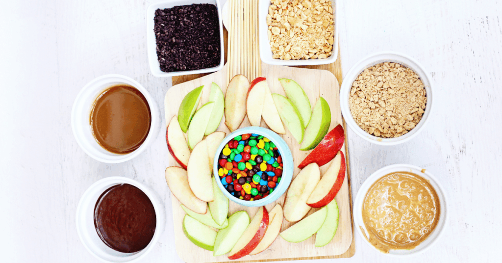 a wooden board with apple slices, m&ms and various toppings