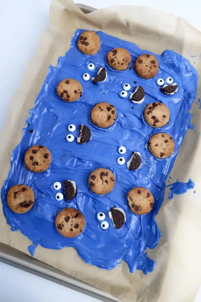 Cookie Monster Bark - Cultivating Brilliant Minds melted blue candy melts spread on a piece of parchment paper with mini cookies and oreos and candy eyes to look like the cookie monster's face