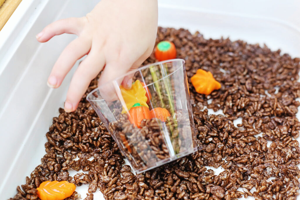 Edible Pumpkin Patch Sensory Bin 7 - Cultivating Brilliant Minds Kid putting the leafs from the Edible Pumpkin Patch Sensory Bin in a clear plastic cup