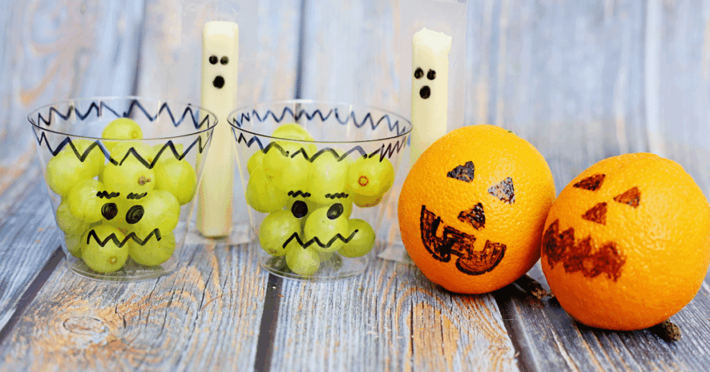 an orange with a jack o lantern face drawn on, a cheese stick with a ghost face, and a cup of green grapes with a frankenstein face
