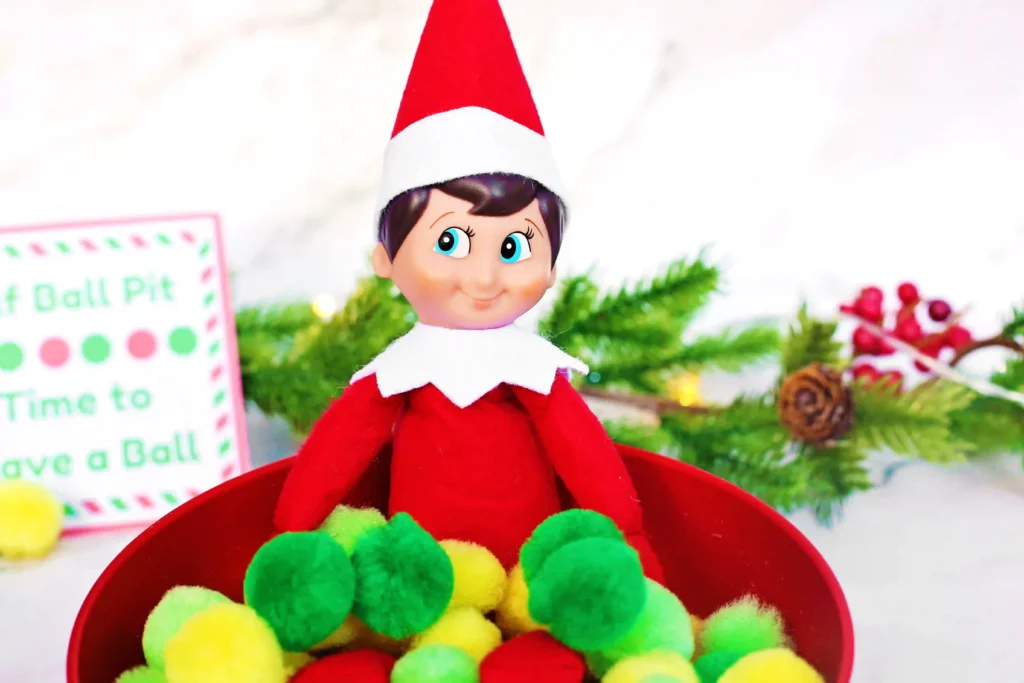 Elf on the Shelf Idea - Cultivating Brilliant Minds elf on the shelf in a bowl of pom poms with a sign in the background that reads "elf ball pit - time to have a ball"