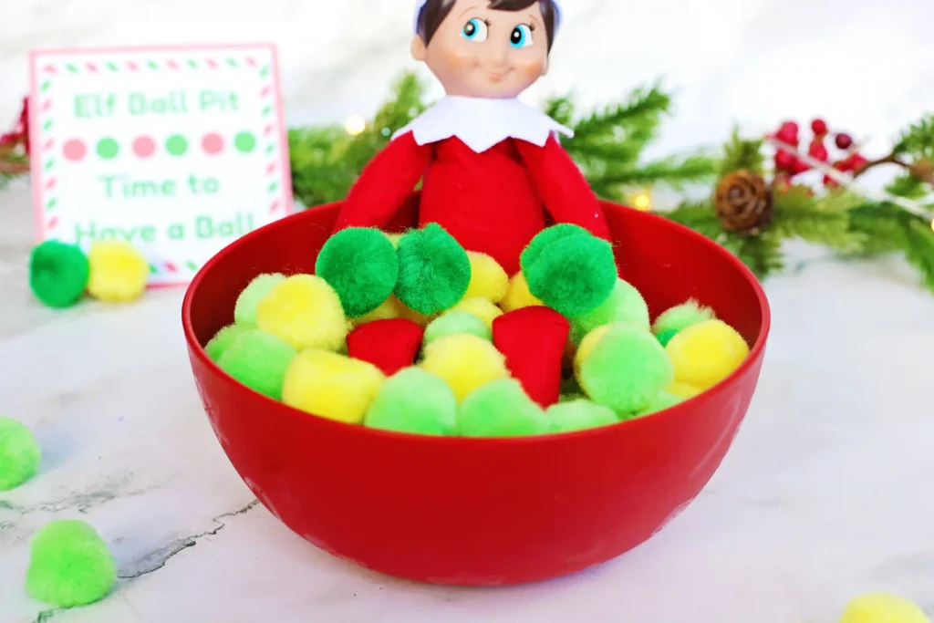 Elf on the Shelf Idea - Cultivating Brilliant Minds elf on the shelf in a bowl of pom poms with a sign that reads "elf ball pit - time to have a ball"