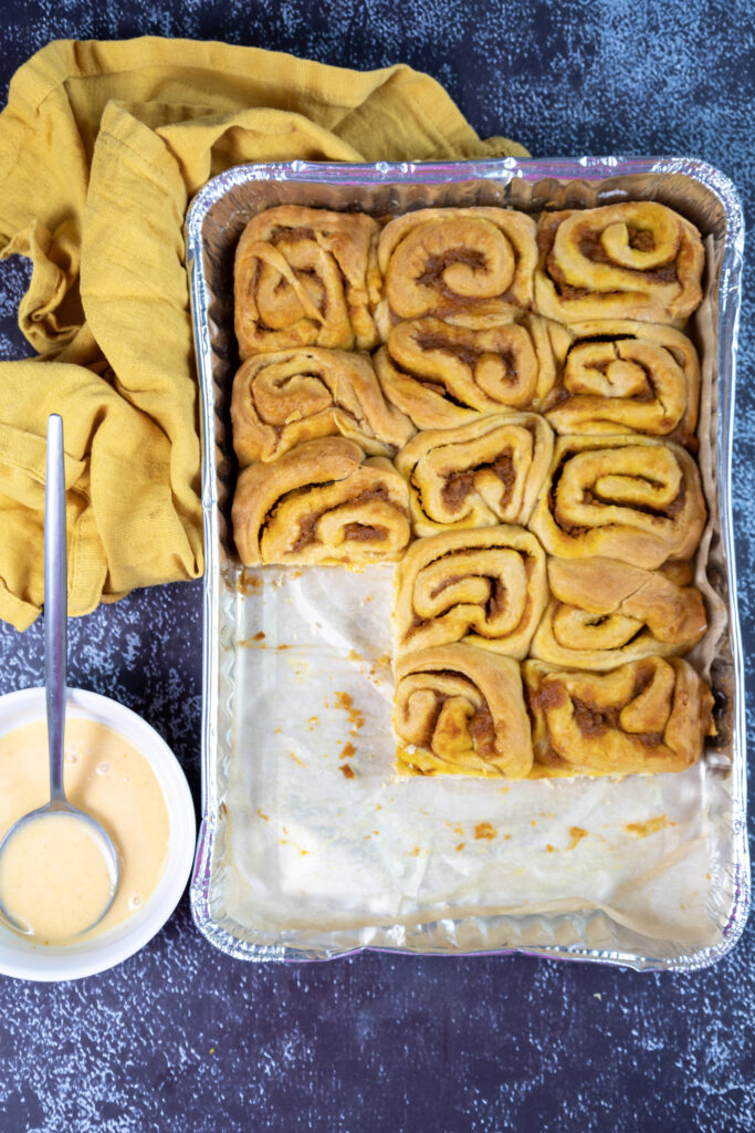 Pumpkin Cinnamon Rolls - Cultivating Brilliant Minds Pumpkin cinnamon rolls in a pan with icing in a bowl