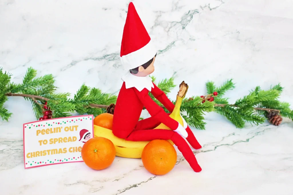 Elf on the Shelf Idea: Banana Races - Cultivating Brilliant Minds an elf on the shelf on a banana made to look like a car with oranges for wheels and a sign that reads "peeling out for christmas cheer"