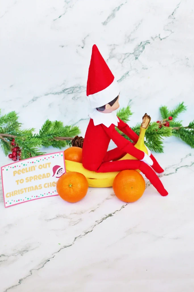 Elf on the Shelf Idea: Banana Races - Cultivating Brilliant Minds an elf on the shelf on a banana made to look like a car with oranges for wheels with a sign that reads "peelin' out to spread christmas cheer"