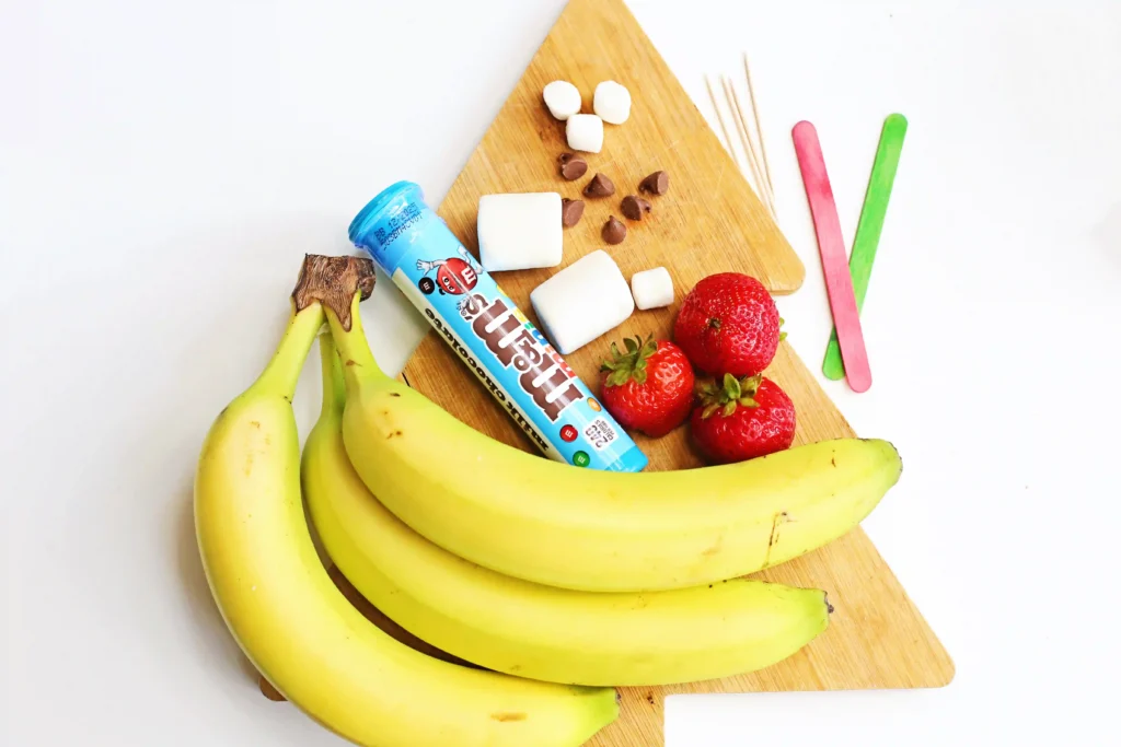 Banana Santa: A Healthy Christmas Snack - Cultivating Brilliant Minds ingredients for the banana santa recipe: bananas, m&ms, strawberries, chocolate chips, marshmallows, toothpicks, and popsicle sticks