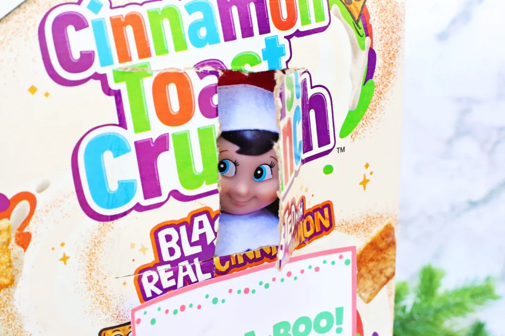 Elf on the Shelf Cereal Box Peekaboo - Cultivating Brilliant Minds a hole cut in a box of cinnamon toast crunch with an elf on the shelf peeking out