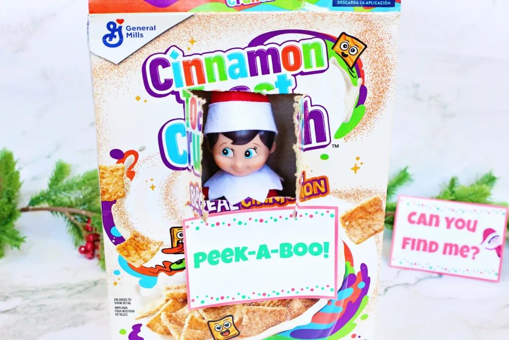 Elf on the Shelf Cereal Box Peekaboo - Cultivating Brilliant Minds a hole cut in a box of cinnamon toast crunch with an elf on the shelf peeking out