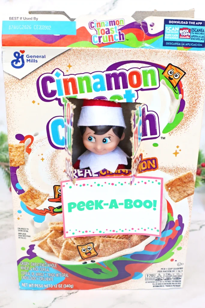 Elf on the Shelf Cereal Box Peekaboo - Cultivating Brilliant Minds a hole cut in a box of cinnamon toast crunch with an elf on the shelf peeking out