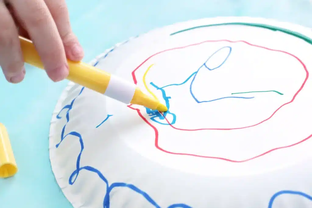 Paper Plate Noisemaker Craft - Cultivating Brilliant Minds a kid drawing with markers on a paper plate
