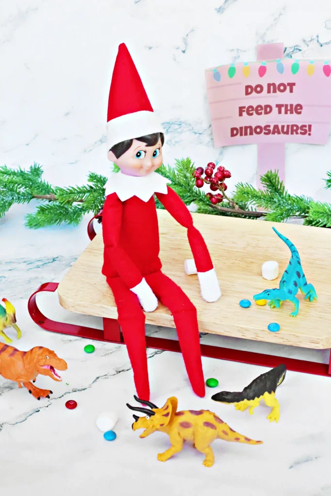Elf on the Shelf Dinosaur Idea - Cultivating Brilliant Minds elf on the shelf sitting on a sled with dinosaurs and candy around him