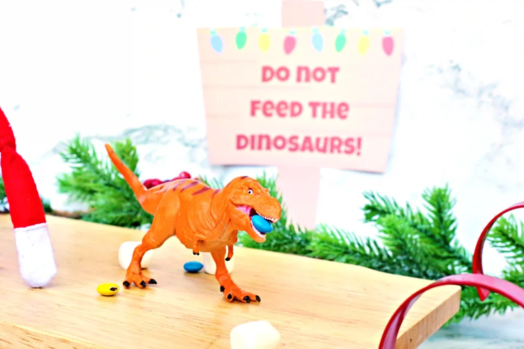 Elf on the Shelf Dinosaur Idea - Cultivating Brilliant Minds elf on the shelf sitting on a sled with dinosaurs and candy around him