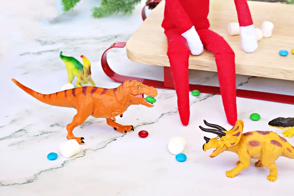 Elf on the Shelf Dinosaur Idea - Cultivating Brilliant Minds elf on the shelf sitting on a sled with dinosaurs and candy around him