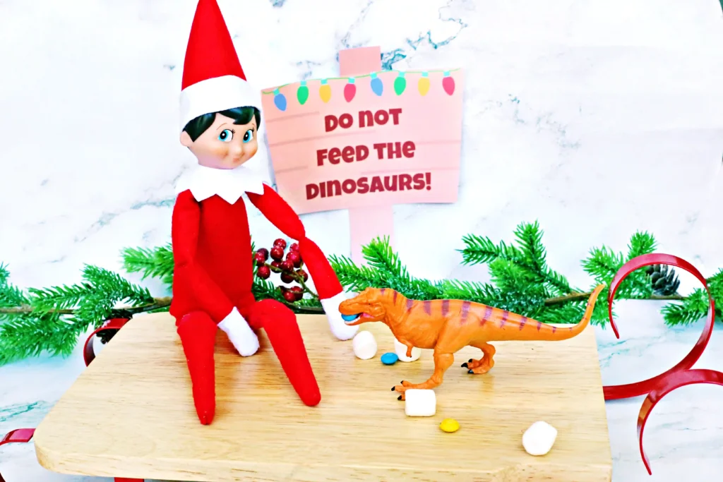 Elf on the Shelf Dinosaur Idea - Cultivating Brilliant Minds elf on the shelf sitting on a sled with dinosaurs and candy around him