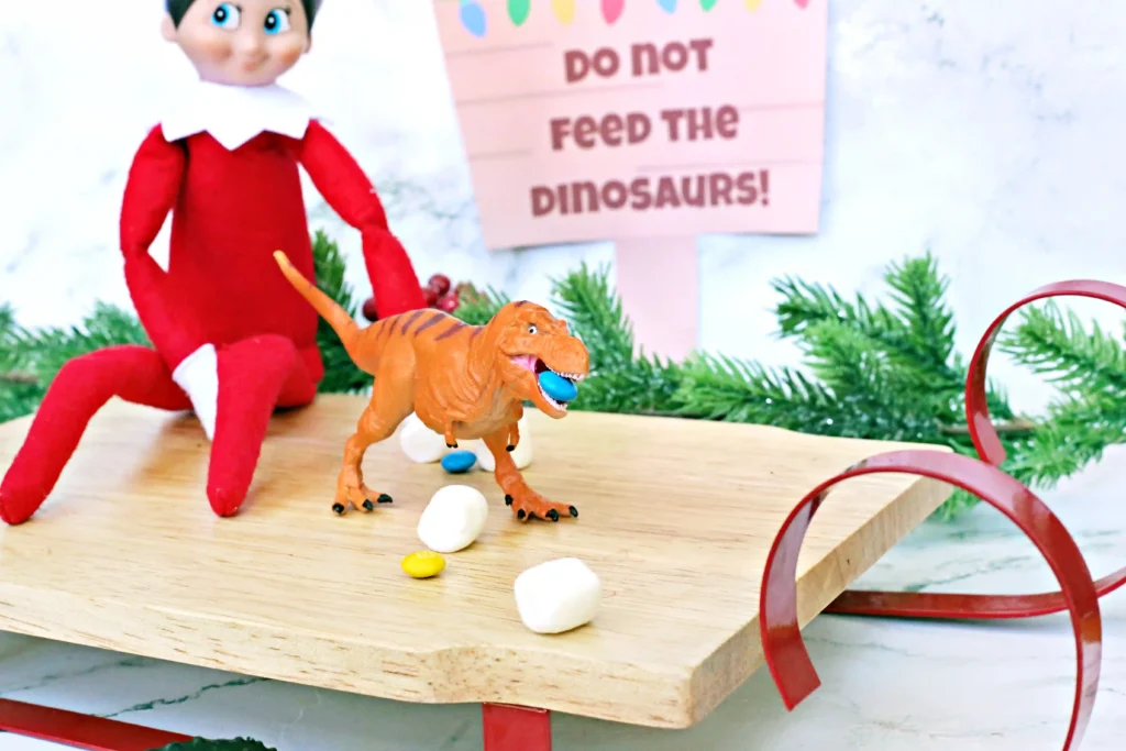 Elf on the Shelf Idea - Cultivating Brilliant Minds elf on the shelf sitting on a sled with dinosaurs and candy around him