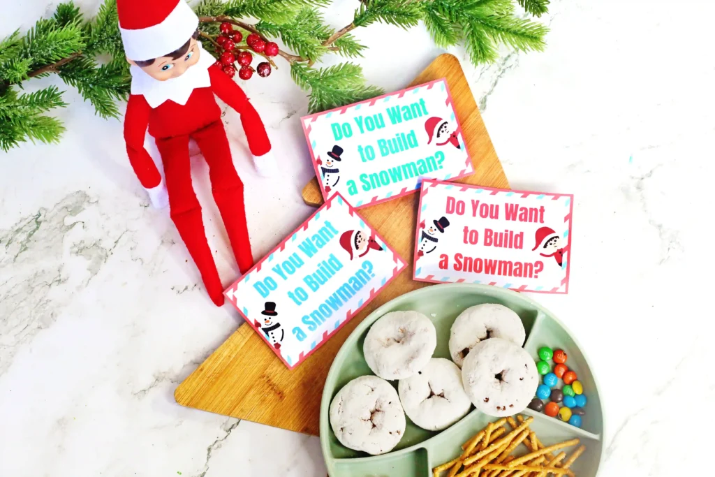 Elf on the Shelf Idea Build A Snowman - Cultivating Brilliant Minds an elf on the shelf next to a tray of donuts, pretzels, m&ms, and a sign that reads "do you want to build a snowman?"