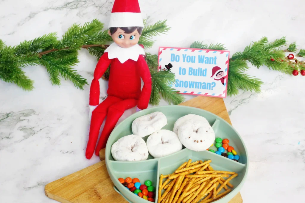 Elf on the Shelf Idea Build A Snowman - Cultivating Brilliant Minds an elf on the shelf next to a tray of donuts, pretzels, m&ms, and a sign that reads "do you want to build a snowman?"