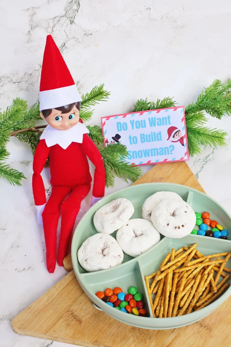 an elf on the shelf next to a tray of donuts, pretzels, m&ms, and a sign that reads "do you want to build a snowman?"