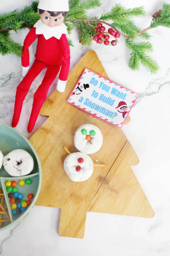 Elf on the Shelf Idea Build A Snowman - Cultivating Brilliant Minds two donuts put together to look like a snowman with pretzel sticks for arms and m&ms for the eyes, nose, and buttons
