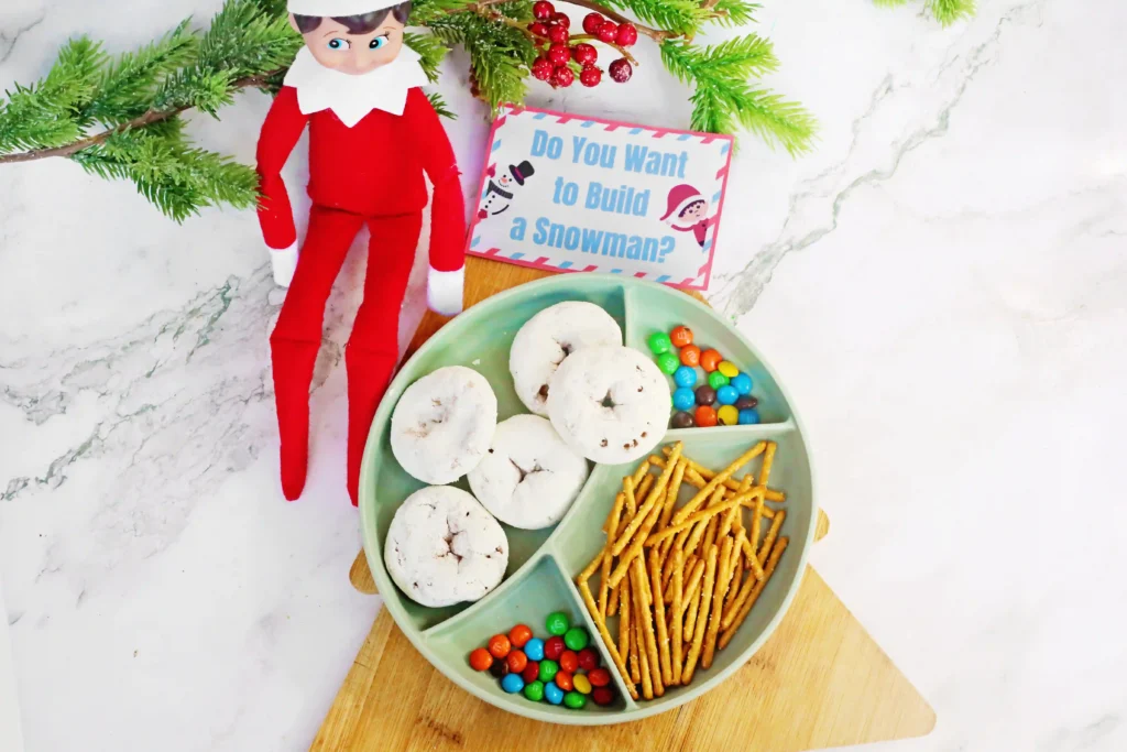 Elf on the Shelf Idea Build A Snowman - Cultivating Brilliant Minds an elf on the shelf next to a tray of donuts, pretzels, m&ms, and a sign that reads "do you want to build a snowman?"