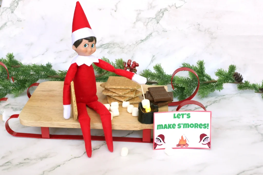 Elf on the Shelf Idea: Elf Campfire - Cultivating Brilliant Minds an elf on the shelf roasting a marshmallow over a flameless candle next to ingredients for s'mores with a sign that reads "let's make s'mores!"