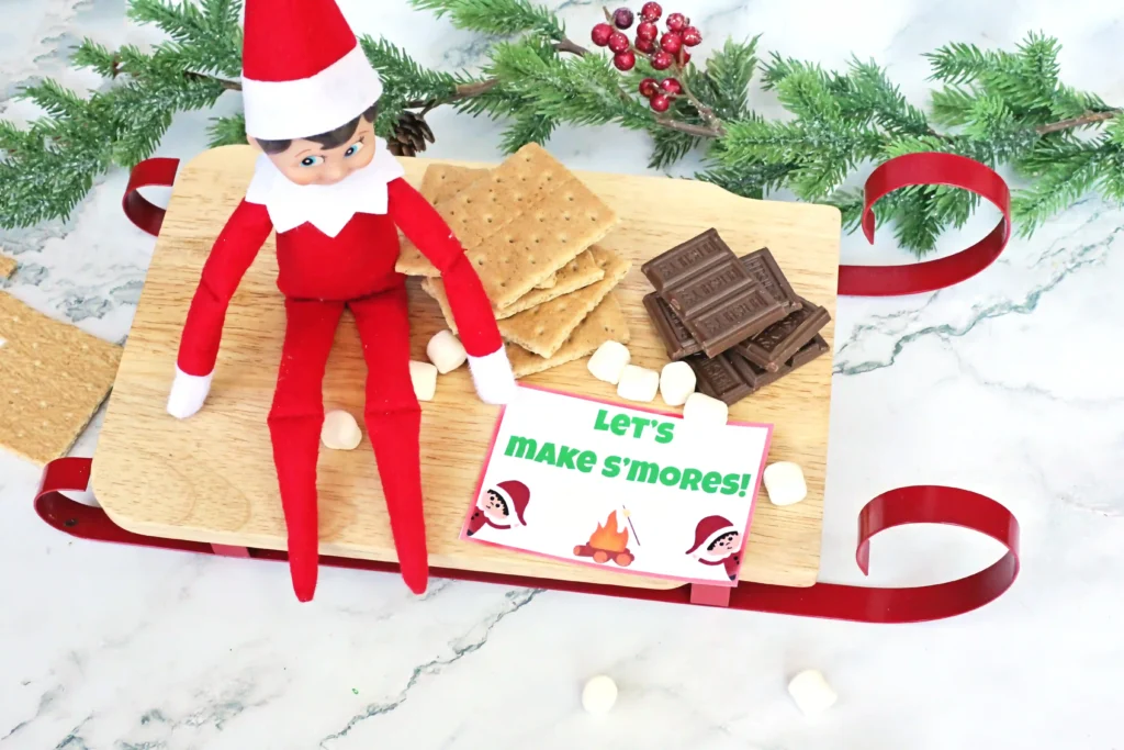 Elf on the Shelf Idea: Elf Campfire - Cultivating Brilliant Minds an elf on the shelf next to ingredients for s'mores with a sign that reads "let's make s'mores!"
