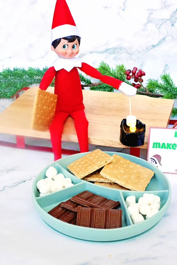 Elf on the Shelf Idea: Elf Campfire - Cultivating Brilliant Minds an elf on the shelf roasting a marshmallow over a flameless candle next to ingredients for s'mores with a sign that reads "let's make s'mores!"