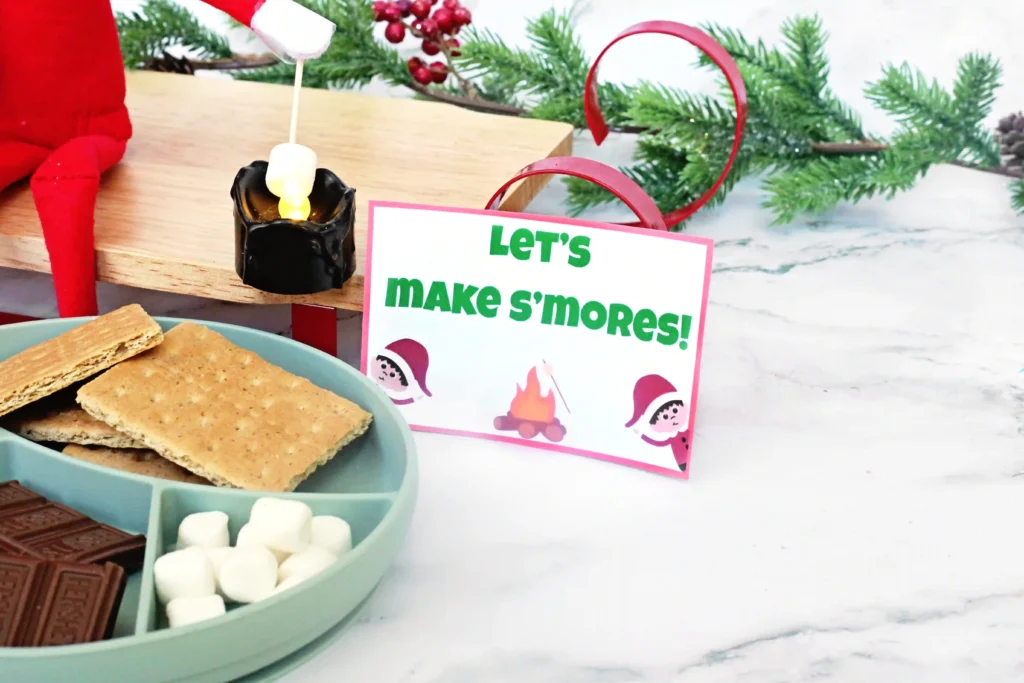 Elf on the Shelf Idea: Elf Campfire - Cultivating Brilliant Minds ingredients for s'mores with a sign that reads "let's make s'mores!"