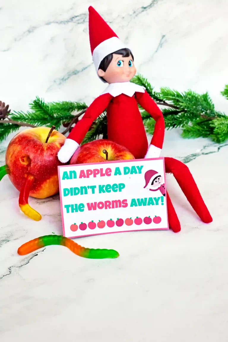 an elf on the shelf next tow apples with gummy worms on them and a sign that reads "an apple a day didn't keep the worms away"