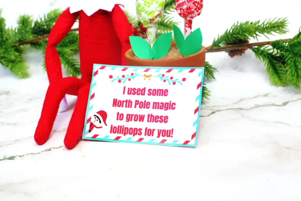 Elf on the Shelf Idea Lollipop Garden - Cultivating Brilliant Minds an elf on the shelf next to a flowerpot filled with lollipops and a sign that reads "I used some north pole magic to grow these lollipops for you!"