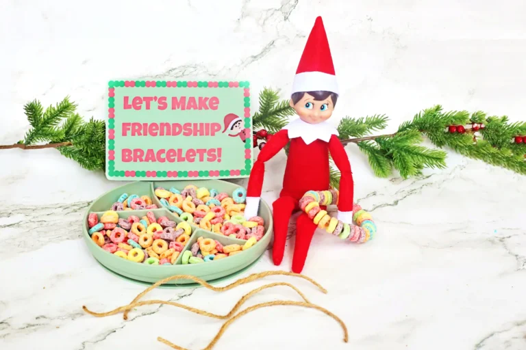 an elf on the shelf holding a fruit loop bracelet next to a bowl of fruit loops and a sign that reads "let's make friendship bracelets"