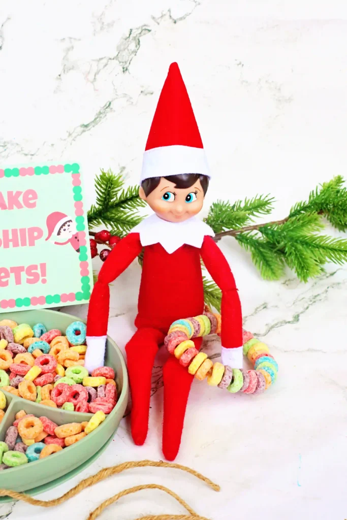 Elf on the Shelf Idea Fruit Loop Bracelets - Cultivating Brilliant Minds an elf on the shelf holding a fruit loop bracelet next to a bowl of fruit loops and a sign that reads "let's make friendship bracelets"