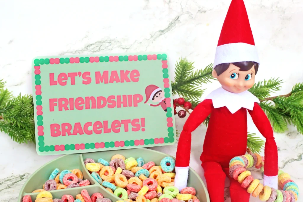 Elf on the Shelf Idea Fruit Loop Bracelets - Cultivating Brilliant Minds an elf on the shelf holding a fruit loop bracelet next to a bowl of fruit loops and a sign that reads "let's make friendship bracelets"