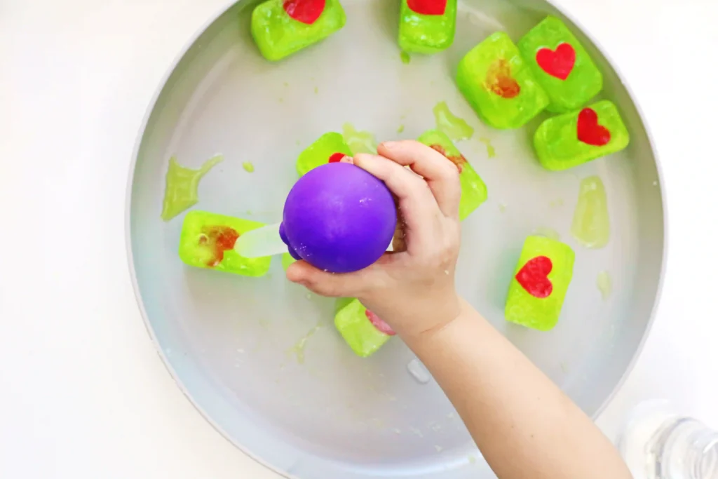 Grinch's Frozen Heart Activity - Cultivating Brilliant Minds a kid holding a pipette over green ice cubes with red hearts inside