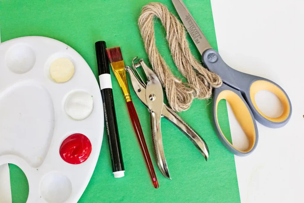 Handprint Santa Ornament - Cultivating Brilliant Minds red, tab, and white paint, a black marker. a paint brush, hole puncher, string, scissors, and green construction paper