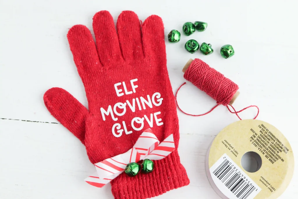 a red glove tied with red and white striped ribbon with green jingle bells that reads "elf moving glove"