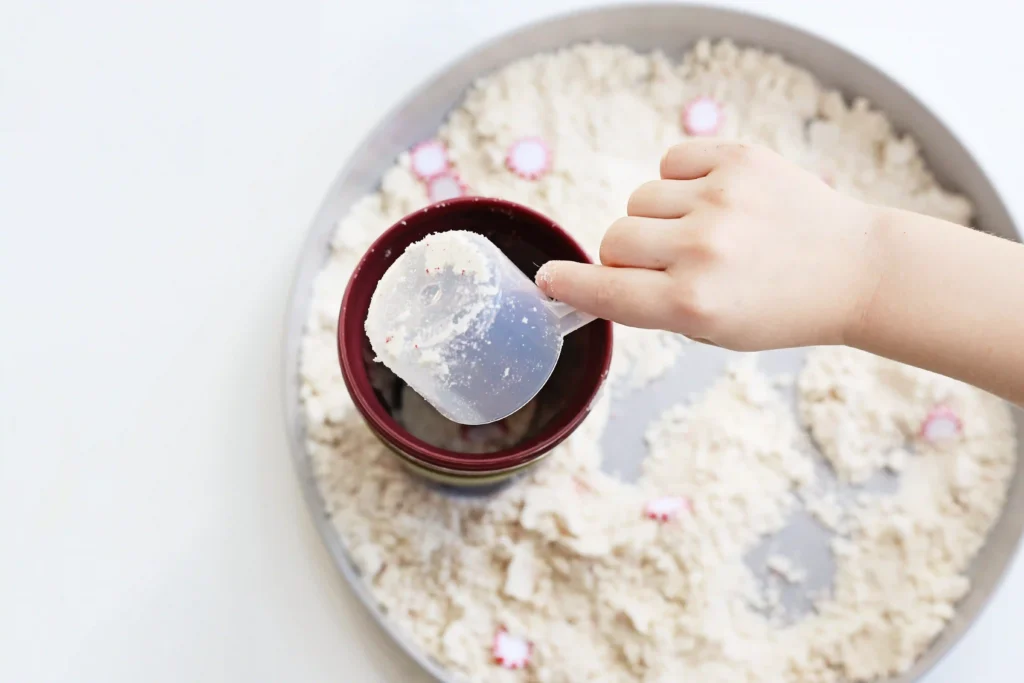 Peppermint Latte Cloud Dough Tray - Cultivating Brilliant Minds a kid scooping up cloud dough from the tray filled with peppermints