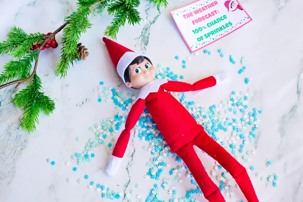 Elf on the Shelf Idea Snow Angel - Cultivating Brilliant Minds an elf on the shelf making a snow angel in sprinkles with a sign that reads "the weather forecast: 100% chance of sprinkles"