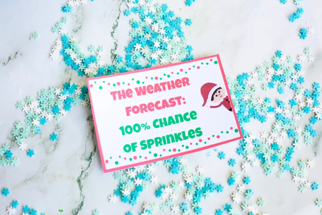 Elf on the Shelf Idea Snow Angel - Cultivating Brilliant Minds an elf on the shelf making a snow angel in sprinkles with a sign that reads "the weather forecast: 100% chance of sprinkles"