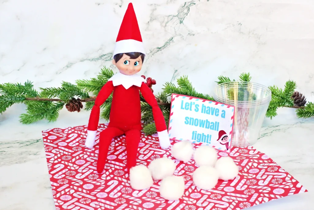 Elf on the Shelf Idea: Snowball Toss Game - Cultivating Brilliant Minds an elf on the shelf with a plastic cup and cotton balls next to a sign that reads "let's have a snowball fight"