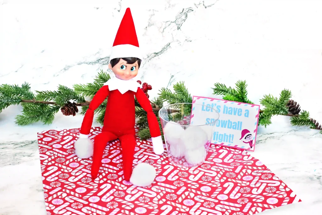 Elf on the Shelf Idea: Snowball Toss Game - Cultivating Brilliant Minds an elf on the shelf with a plastic cup and cotton balls next to a sign that reads "let's have a snowball fight"