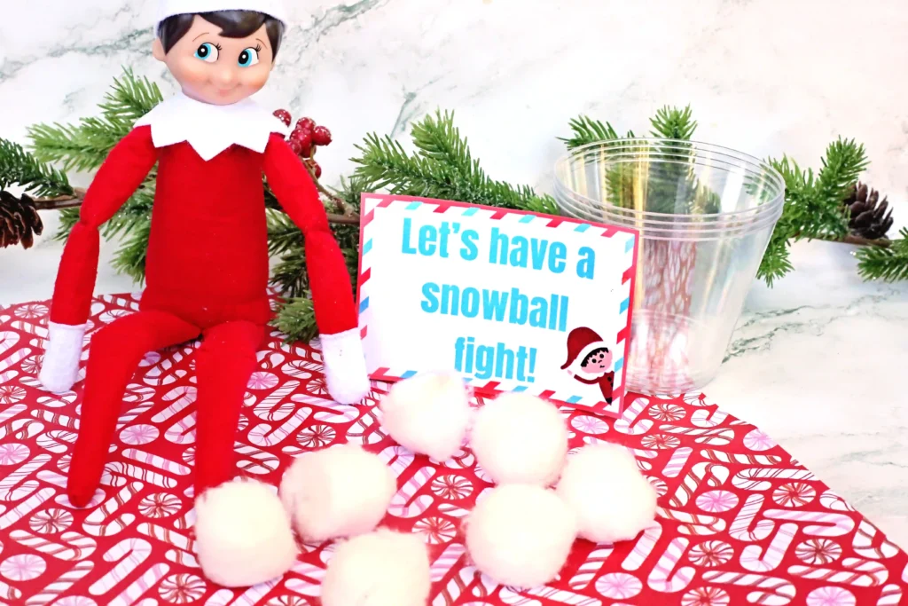 Elf on the Shelf Idea: Snowball Toss Game - Cultivating Brilliant Minds an elf on the shelf with a plastic cup and cotton balls next to a sign that reads "let's have a snowball fight"