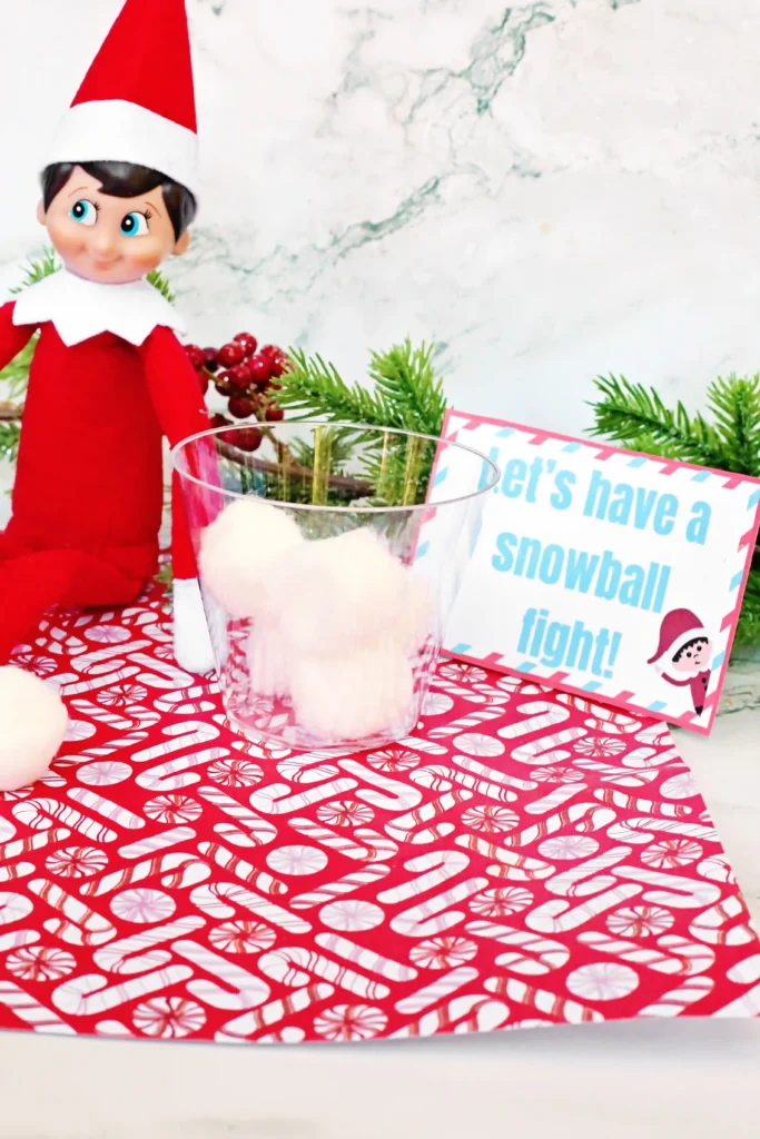 Elf on the Shelf Idea: Snowball Toss Game - Cultivating Brilliant Minds an elf on the shelf with a plastic cup and cotton balls next to a sign that reads "let's have a snowball fight"