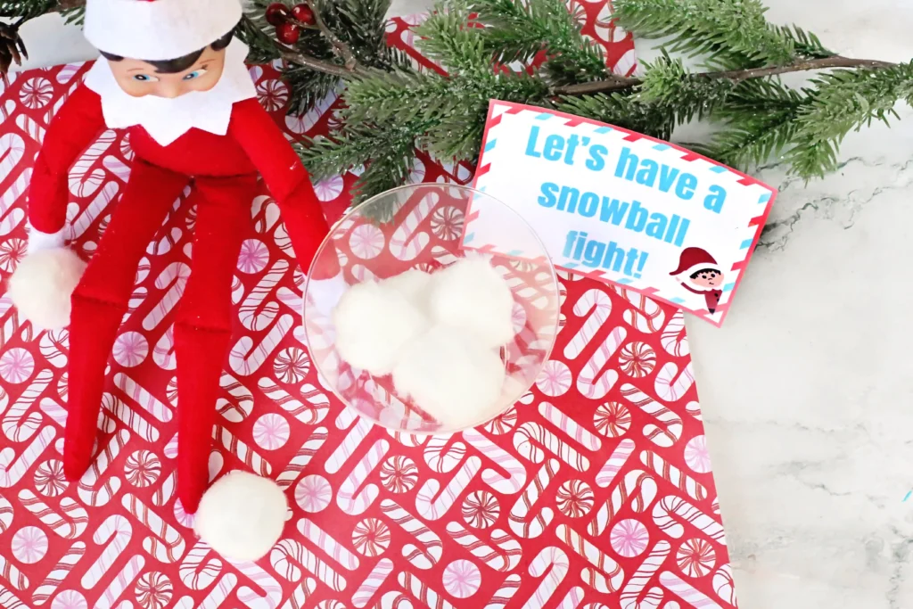 Elf on the Shelf Idea: Snowball Toss Game - Cultivating Brilliant Minds an elf on the shelf with a plastic cup and cotton balls next to a sign that reads "let's have a snowball fight"