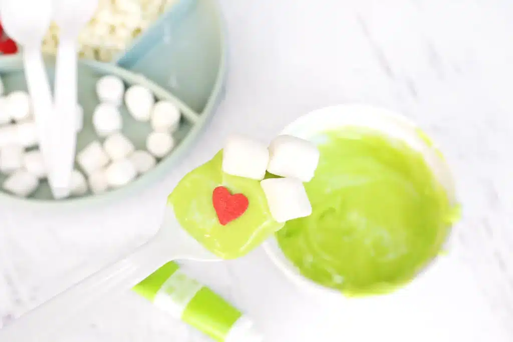 Grinch Hot Chocolate Spoons - Cultivating Brilliant Minds a spoon dipped in melted white chocolate dyed with green food coloring decorated with a red candy heart and mini marshmallows