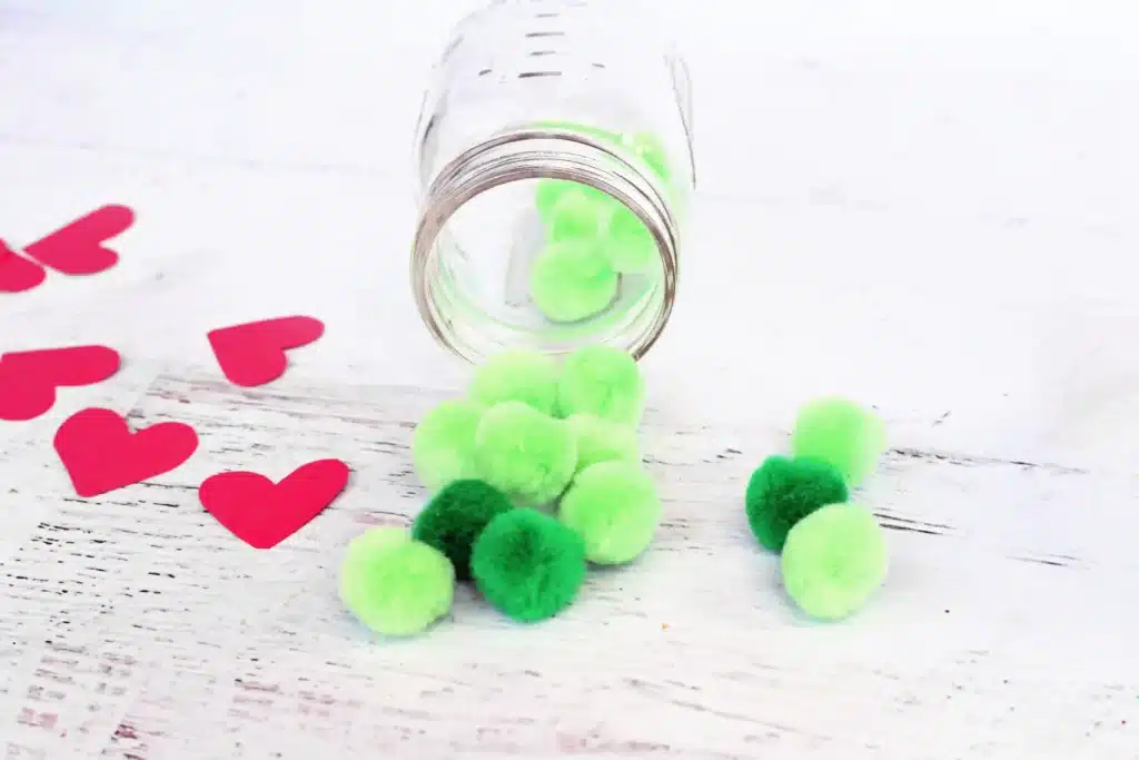 Grinch Heart Jar Challenge - Cultivating Brilliant Minds a mason jar with green pom poms rolling out