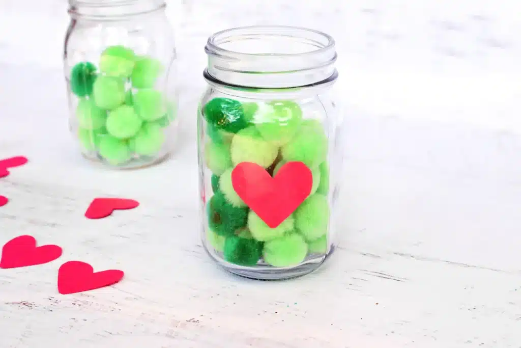 Grinch Heart Jar Challenge - Cultivating Brilliant Minds a mason jar filled with green pom poms and a paper heart taped on