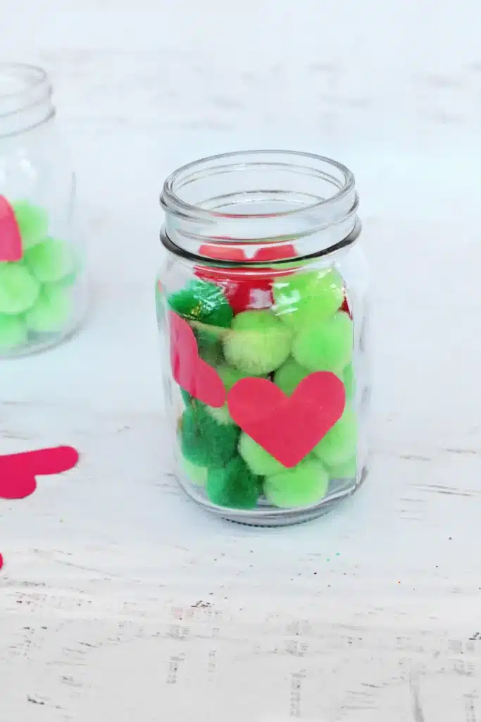 Grinch Heart Jar Challenge - Cultivating Brilliant Minds grinch heart jar challenge: a mason jar filled with green pom poms and a paper heart taped on