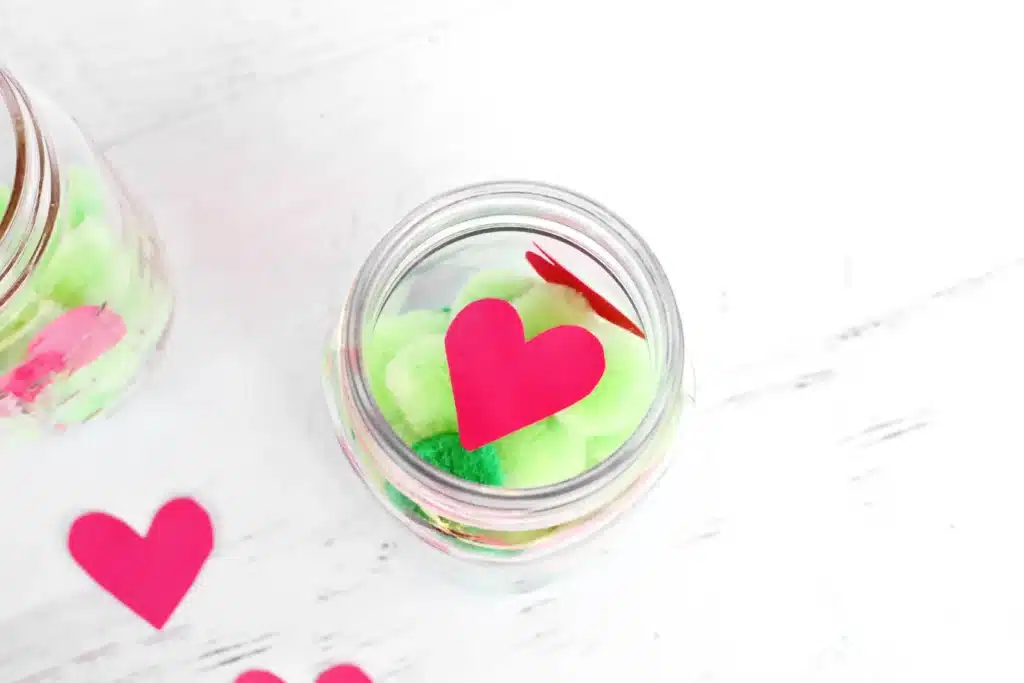 Grinch Heart Jar Challenge - Cultivating Brilliant Minds a mason jar filled with green pom poms and a paper heart taped on
